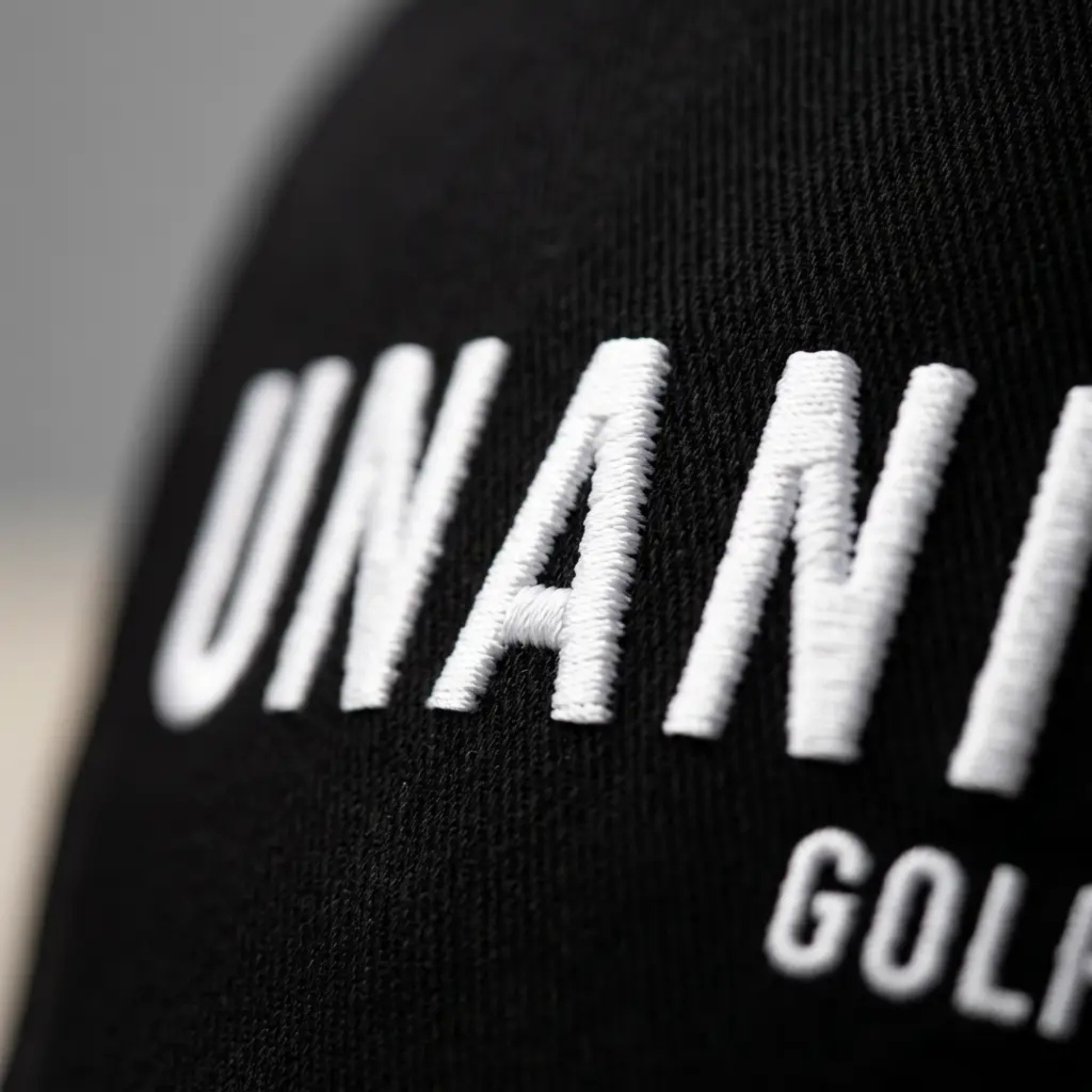 Close-up of black knit fabric with large white embroidered lettering reading 'UNANIMOUS GOLF'.