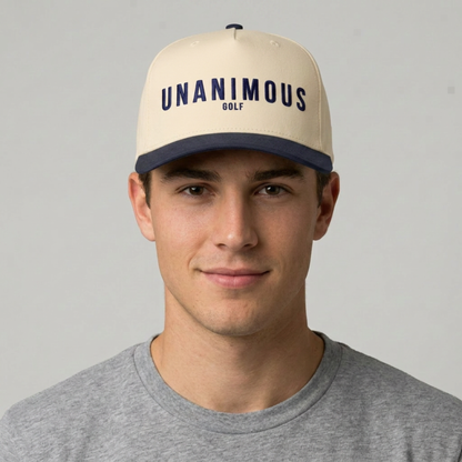 A man wearing a cream-colored baseball cap with a navy blue brim and 'UNANIMOUS GOLF' printed in navy on the front, paired with a gray t-shirt against a light gray background.