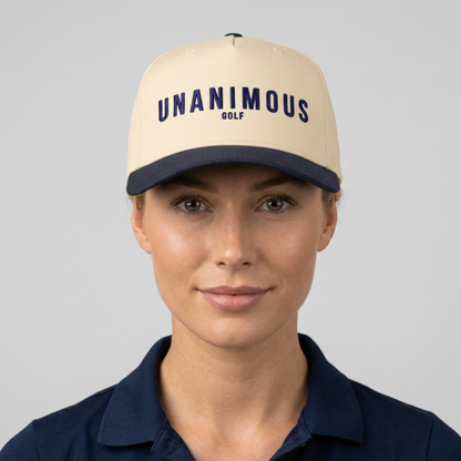 A woman wearing a cream-colored baseball cap with navy blue trim and the text 'UNANIMOUS GOLF' printed on the front, paired with a navy blue polo shirt, against a solid gray background.