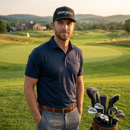A man wearing a navy blue polo shirt, gray trousers, and a black cap with the word 'UNANIMOUS' stands on a golf course with a golf bag beside him.