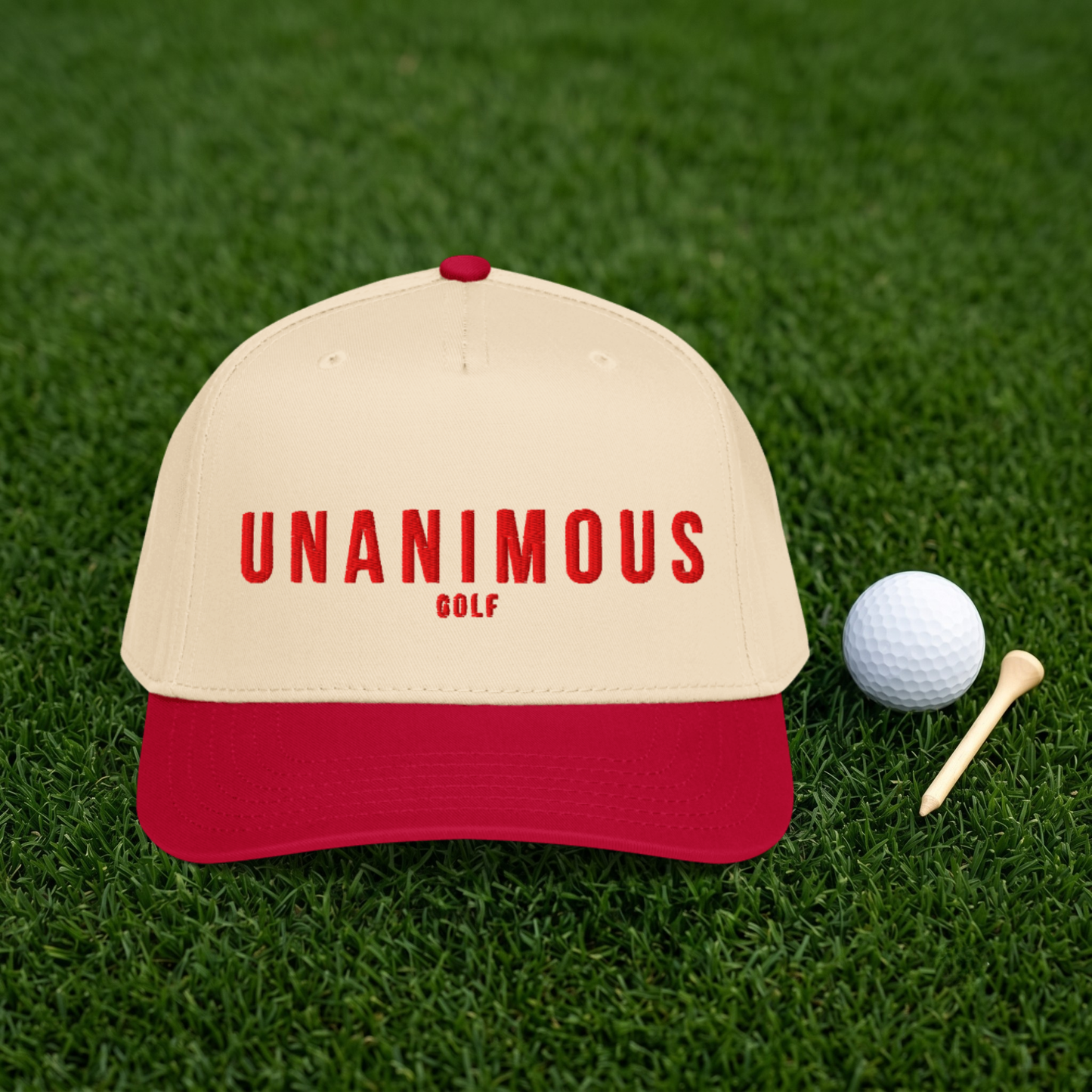 A beige and red baseball cap with 'UNANIMOUS GOLF' printed in red on the front, placed on green grass next to a white golf ball and wooden tees.